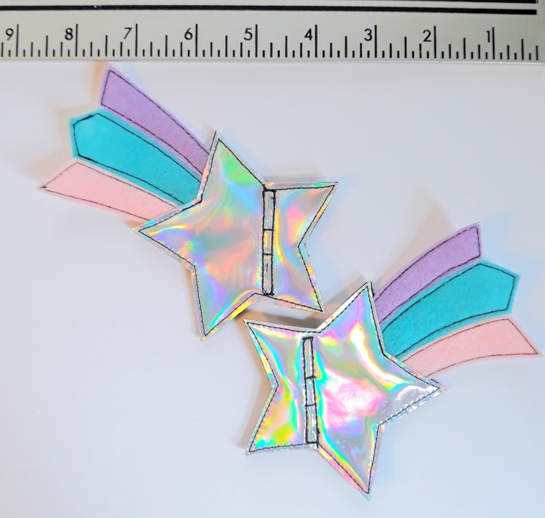 May include: Two iridescent silver star-shaped patches with a rainbow-colored shooting star design. The stars have a black outline and the shooting star is made of pink, blue, and purple felt.