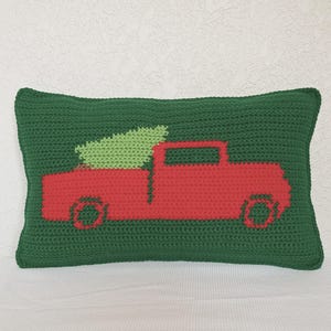 May include: A rectangular, green crocheted pillow featuring a red truck carrying a green Christmas tree. The pillow is made of a textured yarn and is likely handmade. The design is simple and festive, perfect for holiday decor.