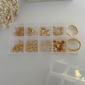 May include: A clear plastic storage box with nine compartments filled with gold-colored jewelry making supplies. The box is open and the compartments are filled with jump rings, earring posts, earring backs, and hoops.