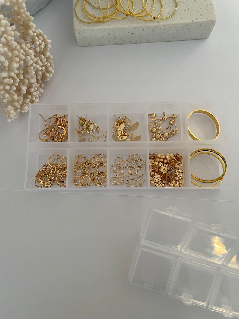 May include: A clear plastic storage box with nine compartments filled with gold-colored jewelry making supplies. The box is open and the compartments are filled with jump rings, earring posts, earring backs, and hoops.