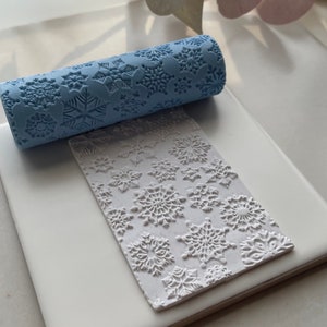 Könnte beinhalten: Eine blaue Teigrolle mit Schneeflockenmuster, die verwendet wird, um geprägte Muster auf Fondant oder Ton zu erstellen. Die Teigrolle liegt neben einer weißen Oberfläche mit einem Schneeflockenmuster.