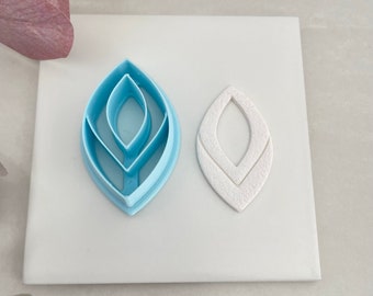 May include: A set of three blue plastic leaf-shaped cookie cutters. The cutters are nested inside each other, with the largest cutter on the bottom and the smallest cutter on top. A white leaf-shaped cookie is shown next to the cutters.
