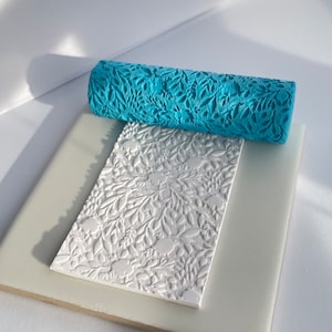 Puede incluir: Un rodillo azul con un diseño floral, utilizado para crear un patrón en relieve en una lámina de fondant blanca.