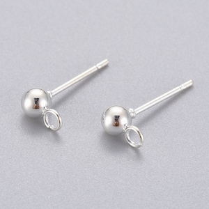 May include: Two silver stud earrings. Each earring has a small, shiny sphere attached to a straight post. A small loop is attached below the sphere. The earrings are shown on a light grey surface.