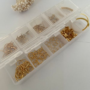 May include: A clear plastic storage box with multiple compartments filled with gold-colored jewelry findings, including jump rings, earring posts, and beads.