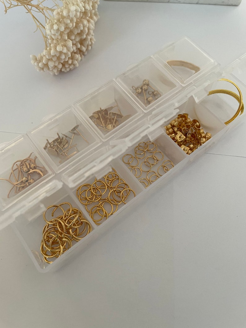 May include: A clear plastic storage box with multiple compartments filled with gold-colored jewelry findings, including jump rings, earring posts, and beads.