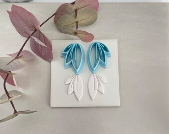 Könnte beinhalten: Ein Set aus Polymer Clay Ausstechern und Prägungen. Die hellblauen Ausstecher sind blattförmig, mit zwei Ausstechern und zwei weißen Blatteindrücken. Das Set ist für das Basteln und Erstellen von blattförmigen Designs konzipiert.