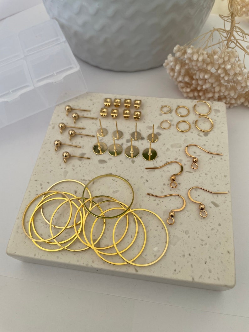 May include: A set of gold-toned earring findings, including hoops, jump rings, earring posts, and earring wires. The findings are arranged on a white marble surface.