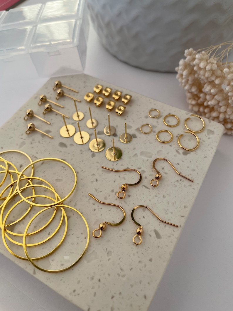 May include: Assortment of gold earring findings, including hoops, hooks, and posts. The findings are arranged on a white marble surface.