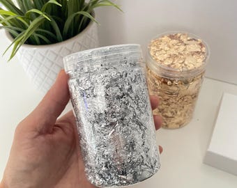 May include: Two clear plastic jars filled with silver and gold leaf flakes. The silver leaf jar is held in a hand. The jars are on a white surface, with a white pot of greenery in the background.