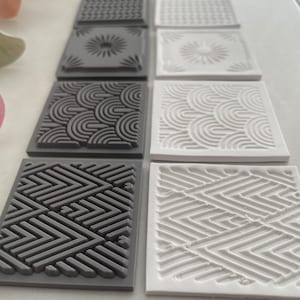 May include: Set of six square silicone stamps with geometric patterns. The stamps are in two colours: grey and white. The patterns include a diamond pattern, a wave pattern, a starburst pattern, and a simple grid pattern.