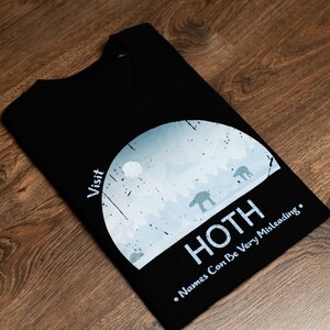 Könnte beinhalten: Schwarzes T-Shirt mit einem Grafikdesign einer schneebedeckten Landschaft mit dem Text "Visit Hoth" und "Names Can Be Very Misleading". Das Design zeigt AT-AT-Walker und einen Mond.