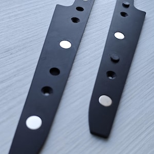 Op de afbeelding: Twee langwerpige, matzwarte onderdelen met een reeks ronde uitsparingen en ingebedde zilverkleurige ronde magneten. De stukken hebben een taps toelopende, hoekige vorm met een getextureerde oppervlakteafwerking, naast elkaar geplaatst.