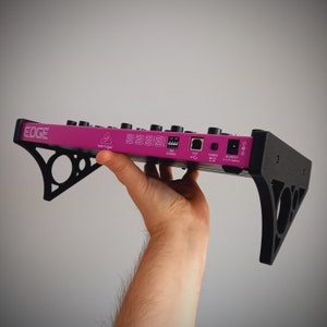 以下が含まれることがあります： 黒い金属製のブラケットが付いたピンクの Behringer EDGE ミキサー。ミキサーの背面パネルには、USB、DC 入力、電源スイッチなどの接続が表示されています。