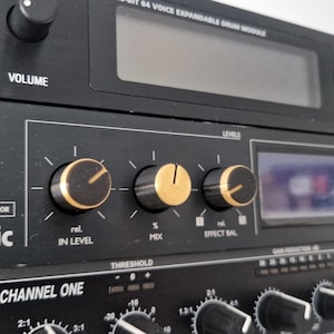 May include: Close-up of a black electronic music mixer with gold knobs. The mixer has a digital display, a volume knob, and a power button. The text "ONE DUAL EFFECTS PROCESSOR" and "electronic" are visible on the front panel.