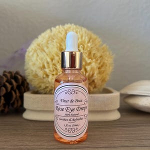 May include: A clear glass bottle of Rose Eye Drops with a white and pink label. The label reads "Fleur de Peau" and "100% Natural". The bottle has a dropper and is 1 fl oz (30ml). A yellow sponge and pine cone are in the background.