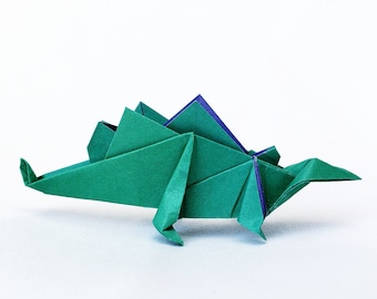 Puede incluir: Dinosaurio de origami verde con detalles morados. El dinosaurio está doblado en un estilo tradicional y tiene una cola larga y una cabeza pequeña.
