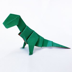 May include: A green origami dinosaur, folded from paper, with a long tail and a small head. The dinosaur is standing on a white surface.