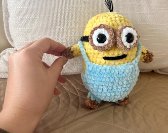 Puede incluir: Un juguete Minion hecho a mano con ganchillo. El Minion es amarillo con ojos marrones, un mechón de pelo negro y un peto azul claro. Los brazos y los pies son marrones. El juguete está sostenido por una mano.