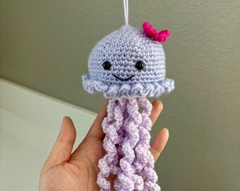 Puede incluir: Una medusa de ganchillo hecha a mano en tonos lavanda y lila claro, con un pequeño lazo rosa. La medusa tiene una cara sonriente y largos tentáculos rizados. Está suspendida por un hilo blanco.