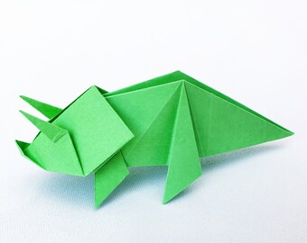 Puede incluir: Un triceratops de origami verde brillante. La escultura de papel presenta una cabeza triangular, tres cuernos y un volante. El dinosaurio está sobre un fondo blanco. Un diseño creativo de arte en papel plegado.