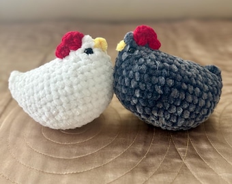 Puede incluir: Dos gallinas de ganchillo, una blanca y otra gris oscuro, están colocadas una al lado de la otra. Cada gallina tiene una cresta roja y un pico amarillo. Las gallinas están hechas de un hilo suave y texturizado.