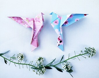 Puede incluir: Dos pájaros de origami hechos de papel con patrones florales. El pájaro de la izquierda es rosa con flores blancas y el pájaro de la derecha es azul con flores blancas. Los pájaros están sentados sobre una superficie blanca con una ramita de flores blancas.