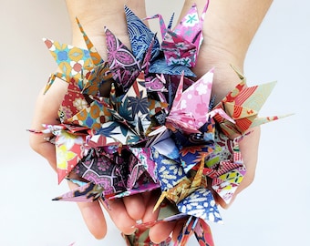 Puede incluir: Una colección de grullas de origami coloridas hechas de papel estampado. Las grullas se sostienen en las manos de una persona.