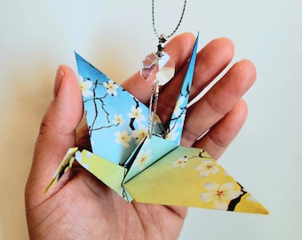 Puede incluir: Una grúa de origami azul y amarilla con un patrón floral cuelga de una cadena plateada con un colgante de cristal.