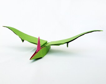 Puede incluir: Pterodáctilo de origami verde con una cresta rosa. La figura de origami está en vuelo con las alas extendidas.