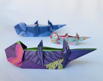 Puede incluir: Tres peces de origami hechos de papel estampado. Los peces están en fila, con el pez más grande en la parte posterior y el pez más pequeño en la parte delantera. El pez más grande es azul con patrones blancos y grises. El pez del medio es azul claro con flores rosas y blancas. El pez más pequeño es morado con patrones verdes, rosas y amarillos.