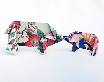Puede incluir: Dos cerdos de origami hechos de papel estampado. El cerdo más grande es verde con flores rosas y blancas, mientras que el cerdo más pequeño es rosa con flores azules y doradas.
