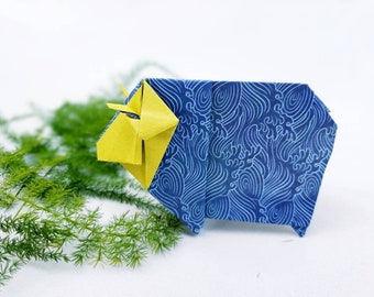 Puede incluir: Una oveja de origami azul con una cabeza amarilla y un patrón de ondas. La oveja está hecha de papel y está sentada sobre una superficie blanca.