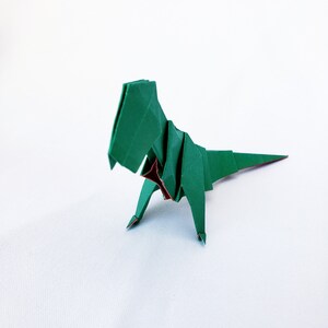 May include: Green origami dinosaur with a red chest, standing on a white surface.