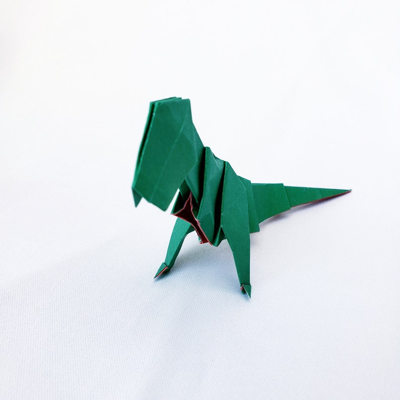 May include: Green origami dinosaur with a red chest, standing on a white surface.