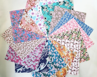 Puede incluir: Una colección de hojas de papel de origami dispuestas en un patrón circular. Las hojas presentan varios patrones florales, incluyendo flores de cerezo, en tonos de rosa, azul, verde y marrón. El papel se muestra sobre una superficie blanca.