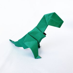 May include: A green origami dinosaur, folded from paper, in a standing position with its head tilted down. The dinosaur has a long tail and a small head.