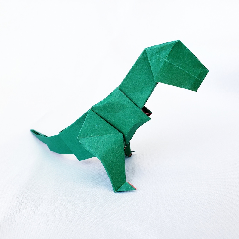 May include: A green origami dinosaur, folded from paper, in a standing position with its head tilted down. The dinosaur has a long tail and a small head.