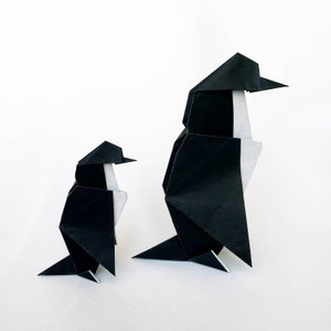 May include: Two origami penguins made of black paper with white accents. The larger penguin is standing upright, while the smaller penguin is standing to the left of the larger penguin.
