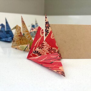 May include: Origami paper cranes in various colors and patterns, including red floral, yellow floral, and blue. A blank brown card stands behind the cranes. The cranes are arranged on a white surface.