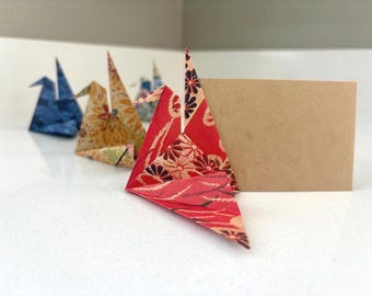 Puede incluir: Grullas de papel de origami en varios colores y patrones, incluyendo floral rojo, floral amarillo y azul. Una tarjeta marrón en blanco se encuentra detrás de las grullas. Las grullas están dispuestas sobre una superficie blanca.