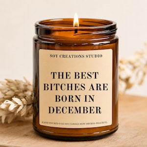Może przedstawiać: Świeca sojowa 255g w bursztynowym szklanym słoiku z beżową etykietą. Etykieta zawiera napis SOY CREATIONS STUDIO, THE BEST BITCHES ARE BORN IN DECEMBER oraz HAND POURED 9 OZ SOY CANDLE-NEW SMYRNA BEACH FL. Widoczny jest płonący knot świecy.