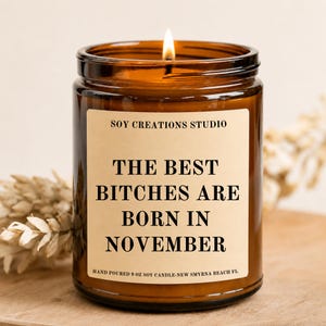 Może przedstawiać: Świeca w bursztynowym szklanym słoiku o pojemności 9 oz z zapalonym knotem i beżową etykietą. Etykieta zawiera czarny tekst: SOY CREATIONS STUDIO, THE BEST BITCHES ARE BORN IN NOVEMBER, oraz HAND POURED 9 OZ SOY CANDLE-NEW SMYRNA BEACH FL.