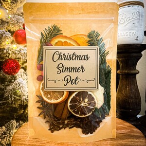 May include: A clear cellophane bag filled with dried orange slices, star anise, cinnamon sticks, cloves, and evergreen sprigs. A label reads "Christmas Simmer Pot". The bag is on a wooden surface, with a Christmas tree and decorative jar in the background.