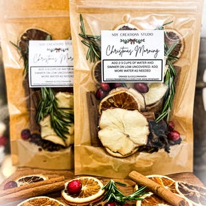 Puede incluir: Dos bolsas transparentes llenas de rodajas de naranja secas, palitos de canela, arándanos y romero. Las bolsas tienen una etiqueta que dice "Christmas Morning" e instrucciones. Los ingredientes están dispuestos sobre una superficie de madera.