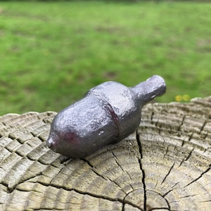 May include: A grey metal acorn-shaped bottle stopper. The stopper is sitting on a wooden surface with a circular grain pattern.