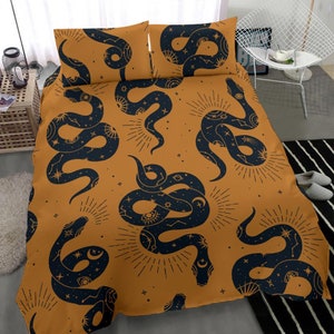 Puede incluir: Un juego de cama de color amarillo mostaza con un patrón de serpiente en blanco y negro. Las serpientes están estilizadas con elementos celestiales como estrellas, lunas y ojos. El juego incluye una funda nórdica y dos fundas de almohada.