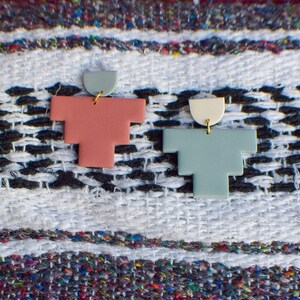 May include: Two geometric earrings. One is a light red-orange color with a light green top, and the other is a light blue-green color with a cream-colored top. Both earrings have a gold-colored hook.
