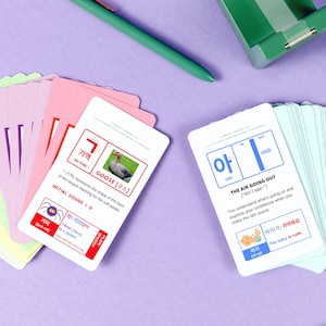 May include: Two flashcards for learning Korean. The first card is pink and features a goose with the Korean character '기역' and the English translation 'GI-YUK'. The second card is blue and features the Korean character '아' and the English translation 'AH'. The card also includes the phrase 'THE AIR GOING OUT' and an illustration of a baby.
