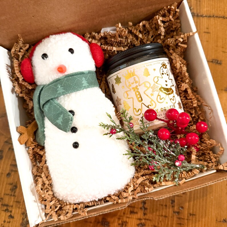 Op de afbeelding: Een wit sneeuwpop knuffel met een groene sjaal en rode oorkleppen, een kleine kaars in een pot met een gingerbread cookie ontwerp, en een takje groen met rode bessen in een bruine kartonnen doos gevuld met gescheurd papier.